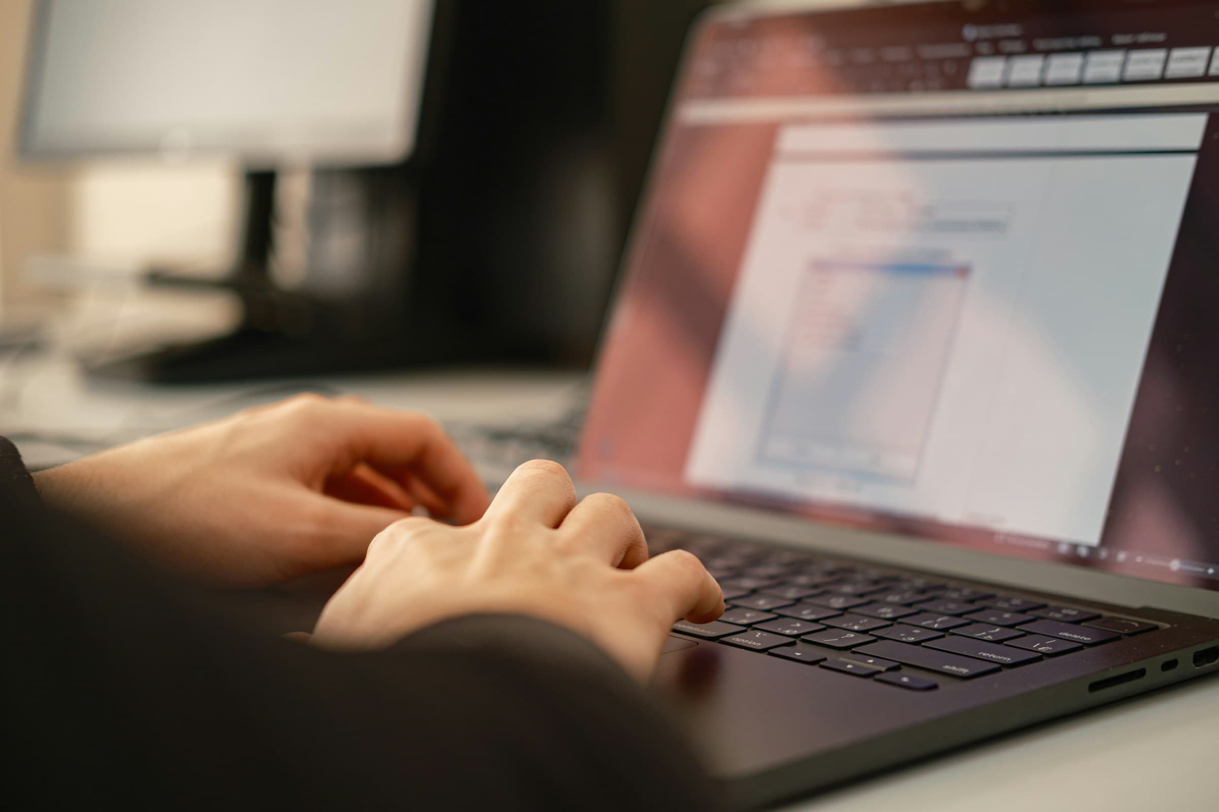 Nahaufnahme zweier Hände, die auf der Tastatur eines Laptops tippen; im Hintergrund unscharf ein externer Monitor.